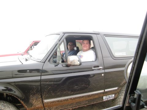 1986 Ford Bronco - Two men are inside a black 1986 Ford Bronco, which has mud on its exterior. A red vehicle is seen in the background.