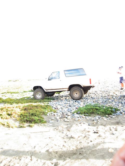 1986 Ford Bronco - White 1980s Ford Bronco parked on a rocky beach with vegetation and a person standing nearby.