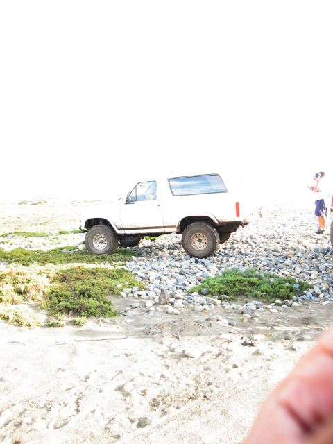 1986 Ford Bronco - A white SUV is parked on a beach with a rocky shoreline and some sparse vegetation.