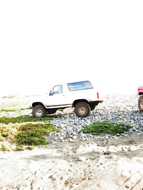 1986 Ford Bronco - A white SUV is parked on a rocky shore next to another vehicle.