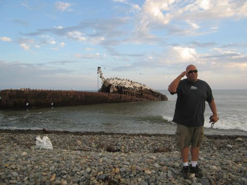 1986 Ford Bronco - A man stands on a pebble beach next to a large, rusted shipwreck partially submerged in the ocean.
