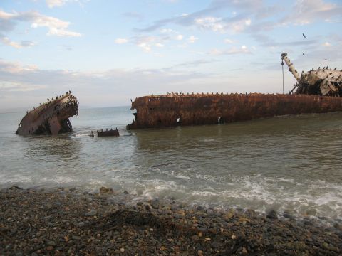 1986 Ford Bronco - Two derelict ships lie partially submerged on a pebble beach under a cloudy sky.