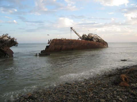 1986 Ford Bronco - Wreckage of a large ship lies partially submerged on a rocky coastline under a cloudy sky.