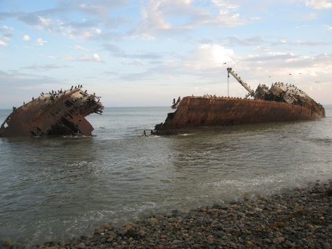 1986 Ford Bronco - A large shipwreck lies partially submerged in the ocean near a rocky coastline under a cloudy sky.