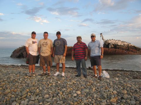 1986 Ford Bronco - A group of men pose for a photo on a pebble beach with a large shipwreck in the background.