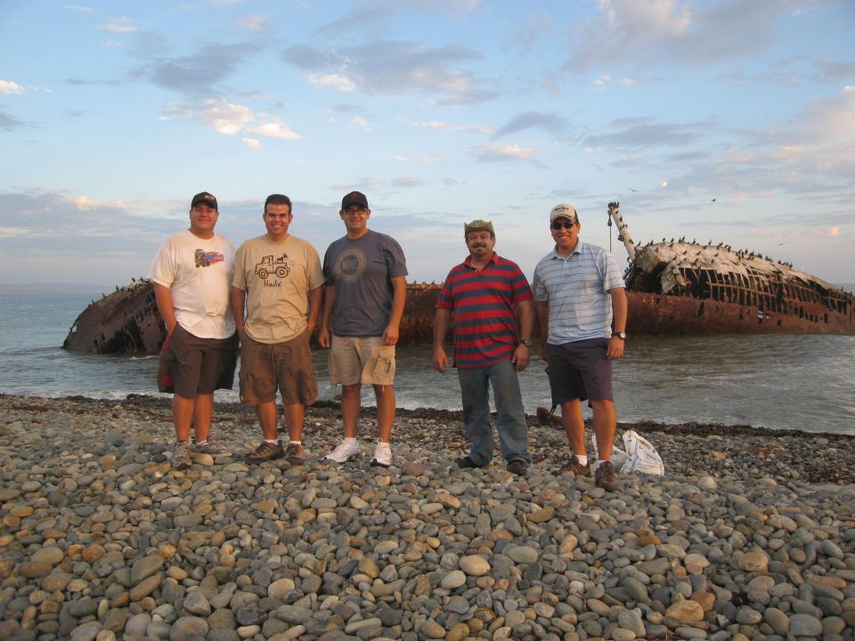 Five men stand on a rocky beach in front of a beached, rusted shipwreck under a cloudy sky