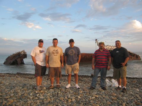 1986 Ford Bronco - Five men pose on a rocky beach with a shipwreck in the background during sunset.