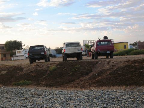 1986 Ford Bronco - Three SUVs are parked on a dirt mound under a cloudy sky. Buildings are visible in the background.