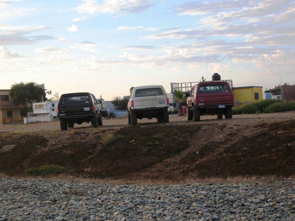 Three SUVs parked on a dirt mound, one black, one white, one red. Buildings and sky in background
