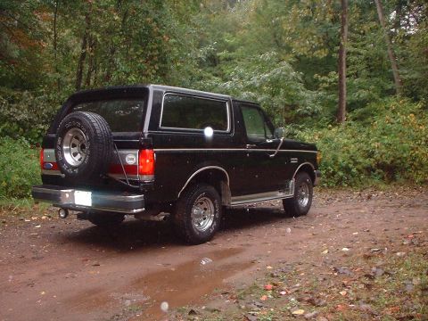1989 Ford Bronco - This black Ford Bronco is parked on a muddy dirt road surrounded by trees.