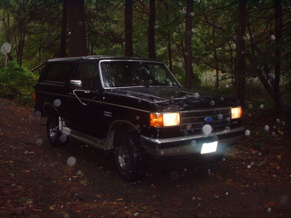 Black 1989 Ford Bronco parked in a forest with rain falling