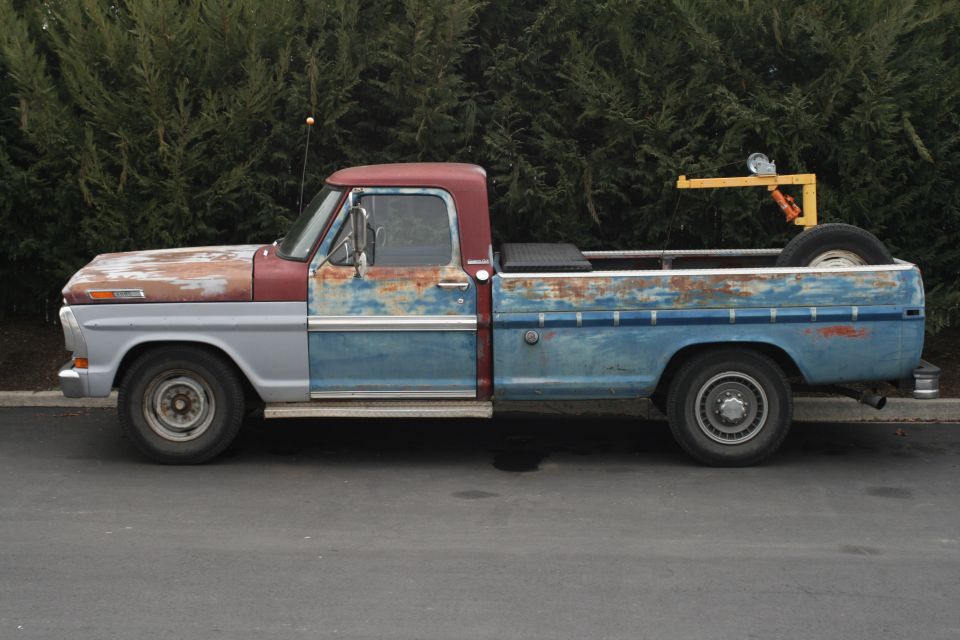 Side view of a beat-up Ford pickup truck with mismatched paint panels and a spare tire mounted in the bed