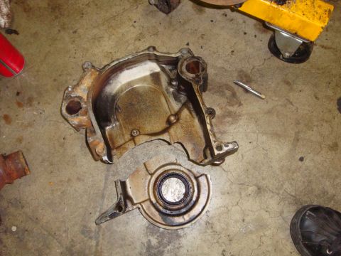 1989 Ford Bronco - Engine timing cover and seal removed from an engine, lying on a concrete floor.