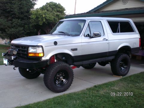 1996 Ford Bronco - Silver and white two-tone Ford Bronco full-size SUV in 3/4 front view, lifted on black steel wheels with mud-terrain tires and red coil-ove…