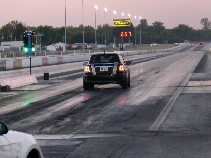 Dark MINI Cooper with white stripes launching at a drag strip at dusk