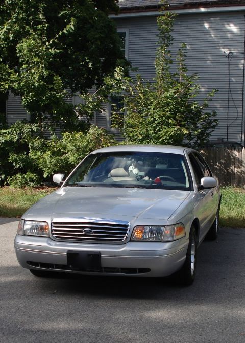 2000 Ford Crown Victoria - A silver 2000 Ford Crown Victoria is parked outside a home, with trees and siding visible in the background.