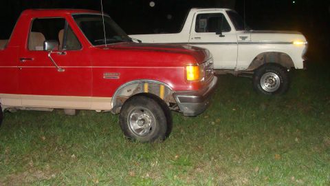 1978 Ford Bronco - Two Ford Broncos, one red and one white, are parked on grass at night.