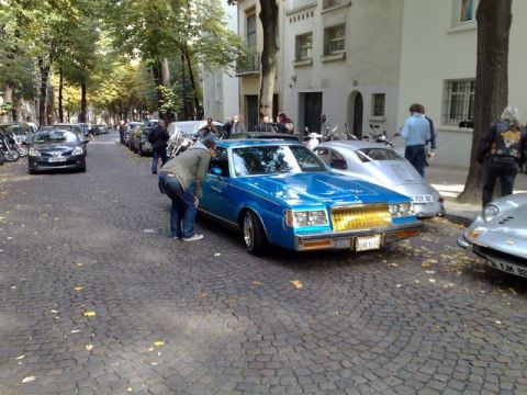 1991 Ford Crown Victoria - A man inspects a bright blue 1980s Buick Regal with a distinctive gold grille, parked on a European-style cobblestone street.