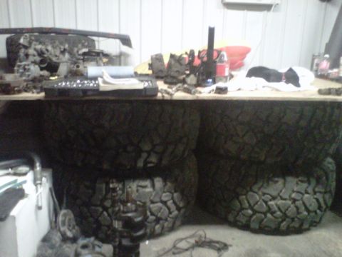 1994 Ford F-150 - A collection of engine components and large off-road tires are stored in a workshop setting.