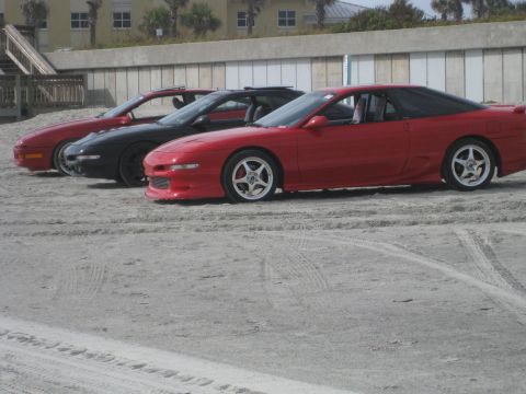 1994 Ford Probe - Three sports cars are lined up on a sandy beach with a building and stairs in the background.