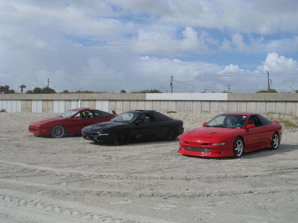 Three 1994 Ford Probes parked on a sandy area with a wall and cloudy sky in the background