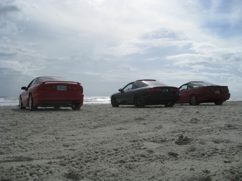1994 Ford Probe - Three coupes are parked on the sand near the ocean under a cloudy sky.