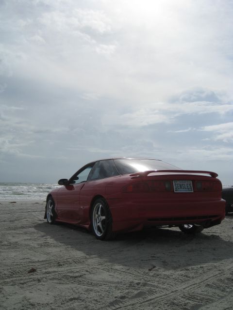 1994 Ford Probe - A red 1994 Ford Probe sits on the sand near the ocean under a cloudy sky.