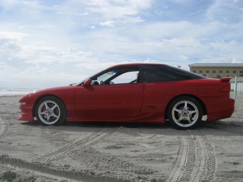 1994 Ford Probe - Side view of a red coupe with a black roof and spoiler on a sandy beach under a cloudy sky.