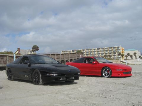 1994 Ford Probe - Two 1994 Ford Probe cars are parked on a sandy beach with a hotel and ocean in the background.