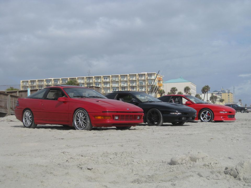 Three Ford Probes parked on a sandy beach with a hotel in the background