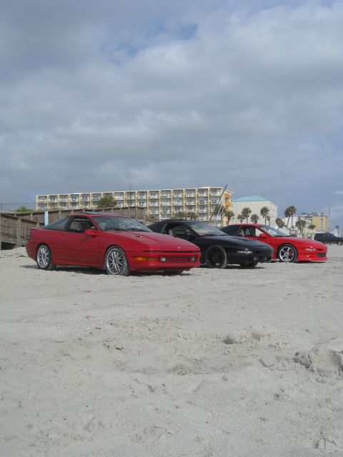 1994 Ford Probe - Three sports cars are parked on a sandy beach with buildings and palm trees in the background.