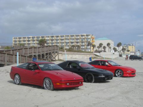 1994 Ford Probe - Three coupes, including a red 1994 Ford Probe, are parked on a sandy beach with a hotel in the background.