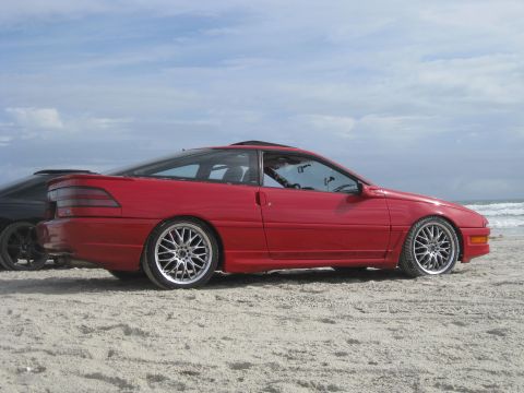 1994 Ford Probe - A red 1994 Ford Probe is seen parked on a beach next to another vehicle, with the ocean waves visible in the distance.