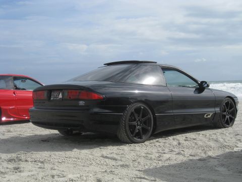 1994 Ford Probe - A black 1994 Ford Probe and a red car sit on a sandy beach near the ocean.