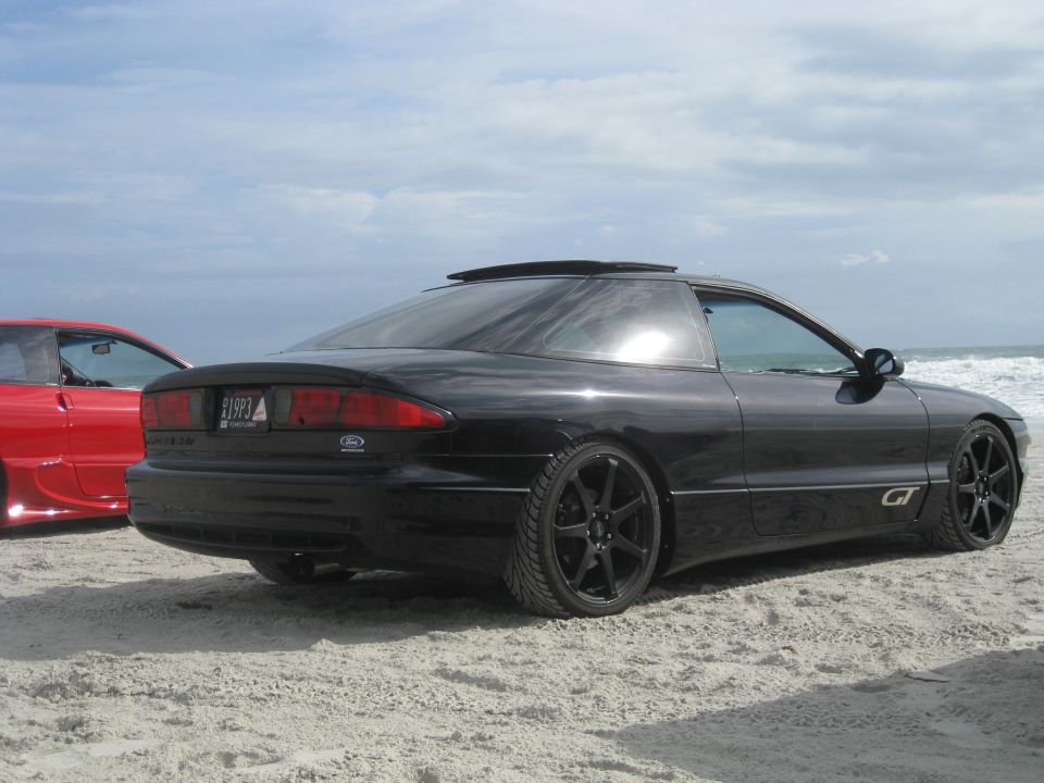 Black 1994 Ford Probe parked on sand with a red car behind it