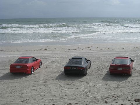 1994 Ford Probe - Three coupes, two red and one black, are parked on a sandy beach with the ocean in the background.