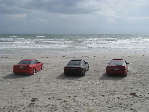 1994 Ford Probe - Three coupes are parked on the sand near the ocean waves under a cloudy sky.
