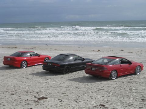 1994 Ford Probe - Three Ford Probe cars are parked on the beach with waves crashing in the background.