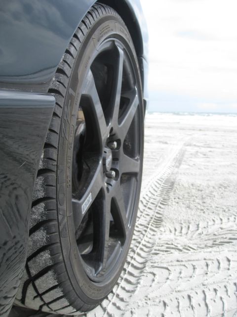 1994 Ford Probe - A close-up view of a car's wheel and tire on a sandy beach, with tire tracks visible in the sand.