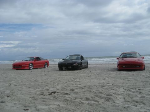 1994 Ford Probe - Three cars are parked on the sand near the ocean under a cloudy sky.