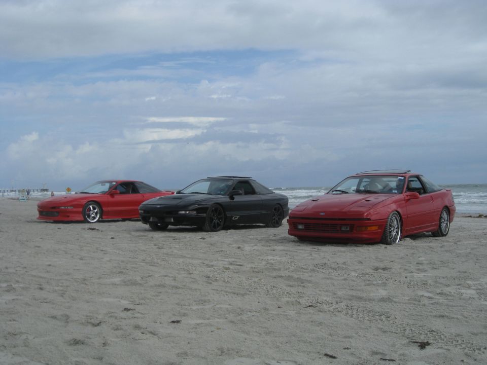 Three red and black Ford Probe cars parked on a sandy beach with the ocean in the background