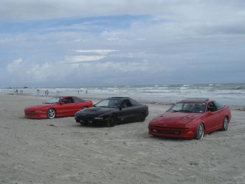 1994 Ford Probe - Three Ford Probes, two red and one black, are parked on a beach under a cloudy sky.