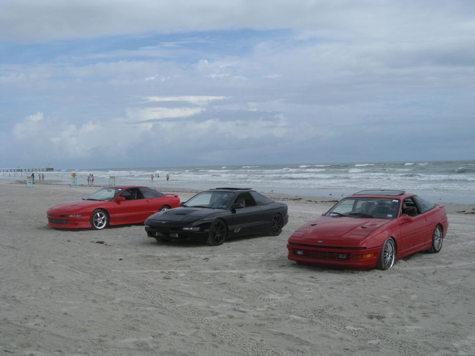 Three red and black 1994 Ford Probes parked on a sandy beach with the ocean in the background