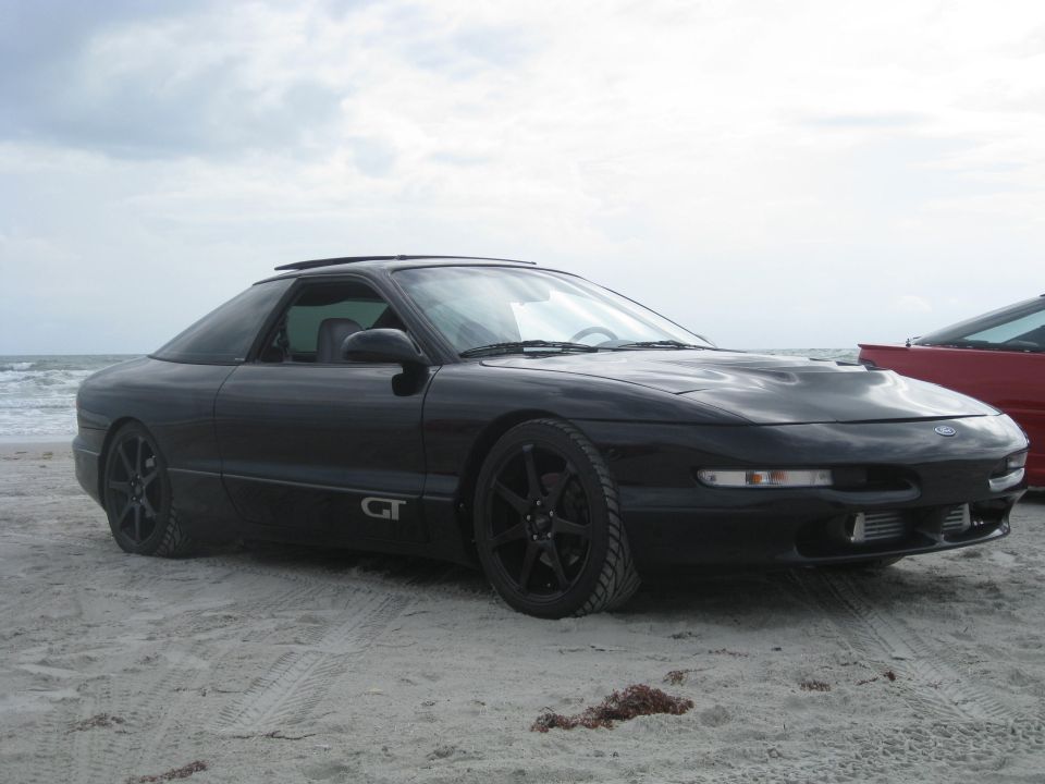 Black Ford Probe GT parked on a sandy beach with the ocean in the background. Another red car is partially visible to the right