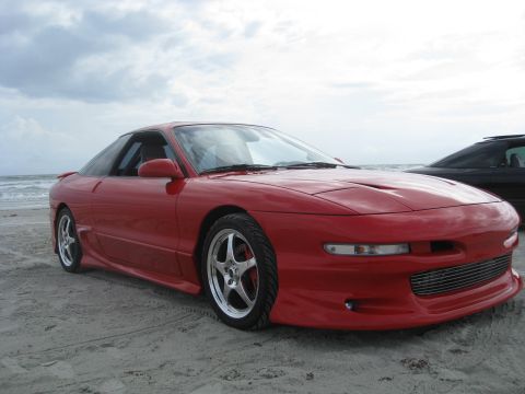 1994 Ford Probe - A red sports coupe sits on the sand near the ocean, with another dark vehicle partially visible behind it.