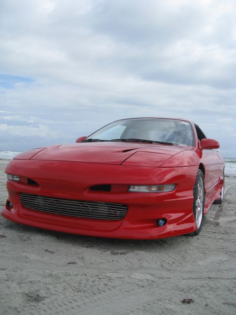 1994 Ford Probe - A red sports car sits on the sand near the ocean under a cloudy sky.