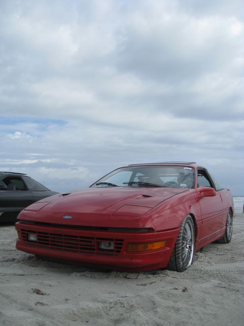 1994 Ford Probe - A red coupe with pop-up headlights sits on a sandy beach next to another vehicle under a cloudy sky.