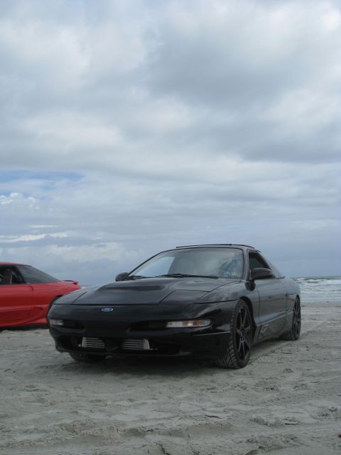 1994 Ford Probe - A black 1994 Ford Probe sits on the sand near the ocean, with another vehicle visible behind it under a cloudy sky.