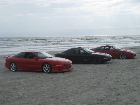 1994 Ford Probe - Three Ford Probes are parked on the beach near the ocean waves under a cloudy sky.