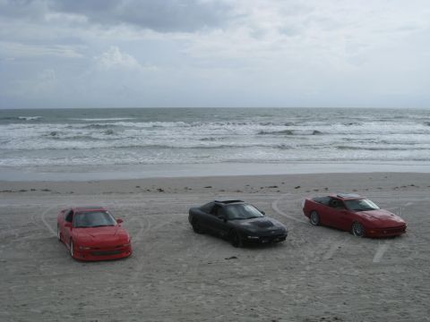 1994 Ford Probe - Three cars are parked on the sand near the ocean waves under a cloudy sky.